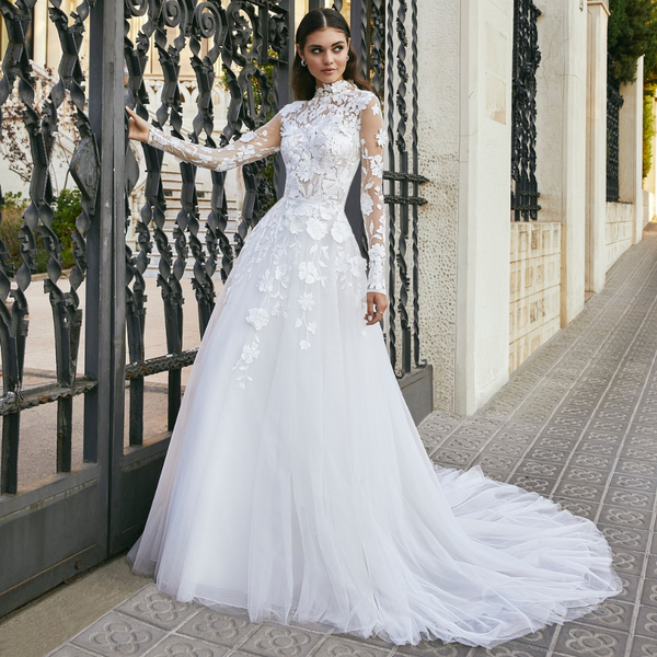 Model stood by black gate in Ronald Joyce style 69717, a high neck A-line wedding dress with lace neckline and sleeves, 3D flowers and a tulle skirt and train. 